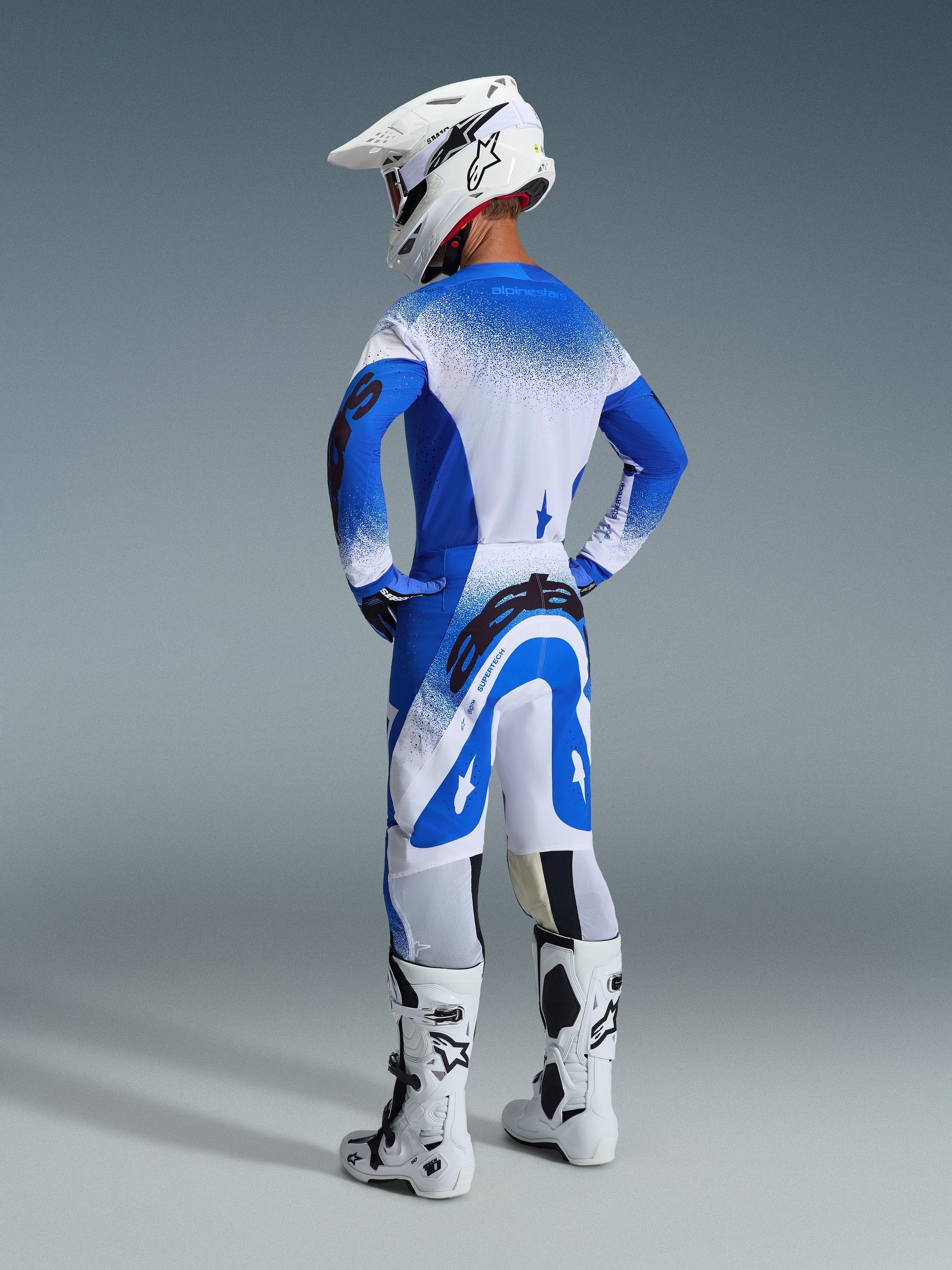 Alpinestars 2026 Supertech Scenz Gear Set, back view of a rider wearing the Ucla Blue White jersey and pants, white motocross helmet, and white racing boots, pro-level racing performance outfit