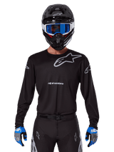Alpinestars 2025 Racer Graphite Jersey, Motocross Racing Gear, Black with Gray logos, worn with matching black helmet, blue goggles, and blue and gray gloves, form-fitting performance design with chest and arm branding