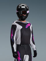 Motocross rider wearing the Alpinestars 2026 Fluid Grid Jersey, Black, Light Gray, and Purple, featuring large pink and purple star logos, paired with matching pants, black helmet, and goggles, complete off-road racing gear set