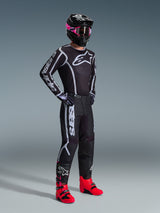 Motocross rider wearing 2026 Fluid Apex Jersey, Black and Gray, paired with matching pants, black helmet with pink goggles, and bright red boots, featuring bold white Alpinestars branding on the chest and sleeves, coordinated professional off-road racing outfit.