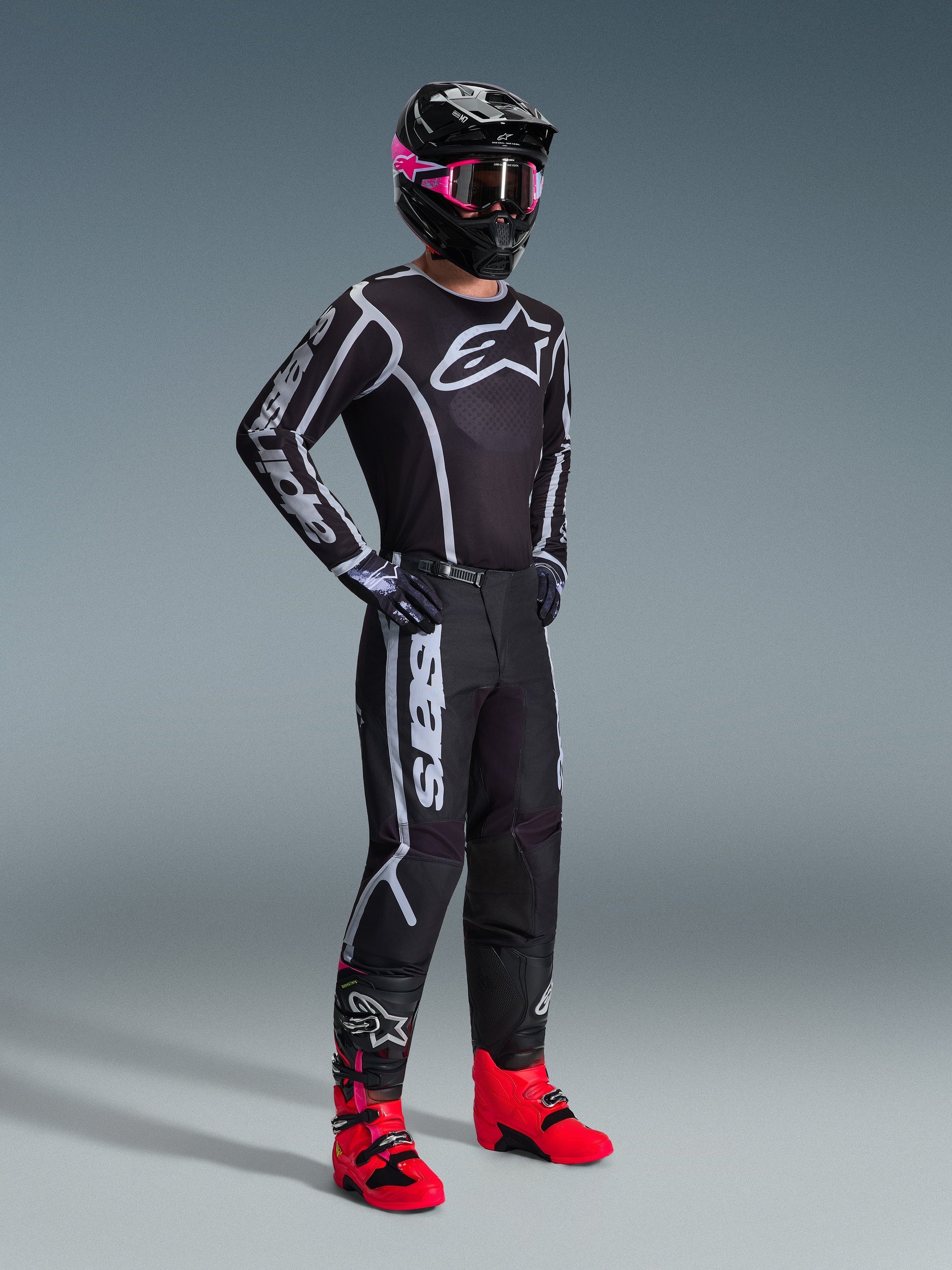 Motocross rider wearing 2026 Fluid Apex Jersey, Black and Gray, paired with matching pants, black helmet with pink goggles, and bright red boots, featuring bold white Alpinestars branding on the chest and sleeves, coordinated professional off-road racing outfit.