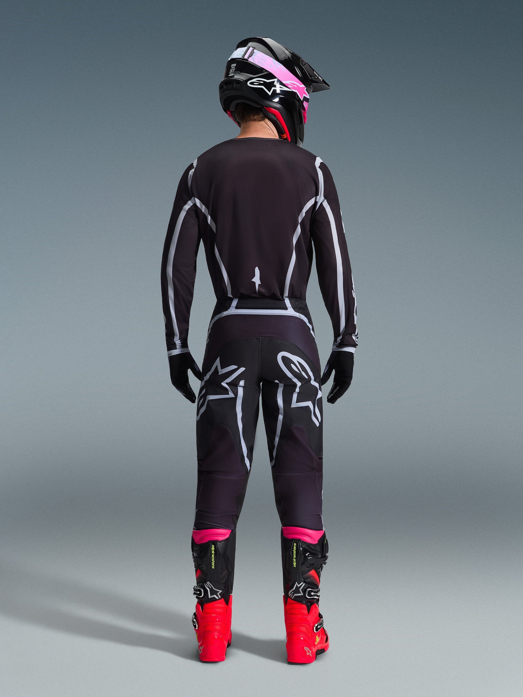 Motocross rider from the back wearing the Alpinestars 2026 Fluid Apex Jersey and matching pants in Black/Gray, featuring white geometric design lines and star logos on the seat, accessorized with a black and pink helmet and red and black racing boots.