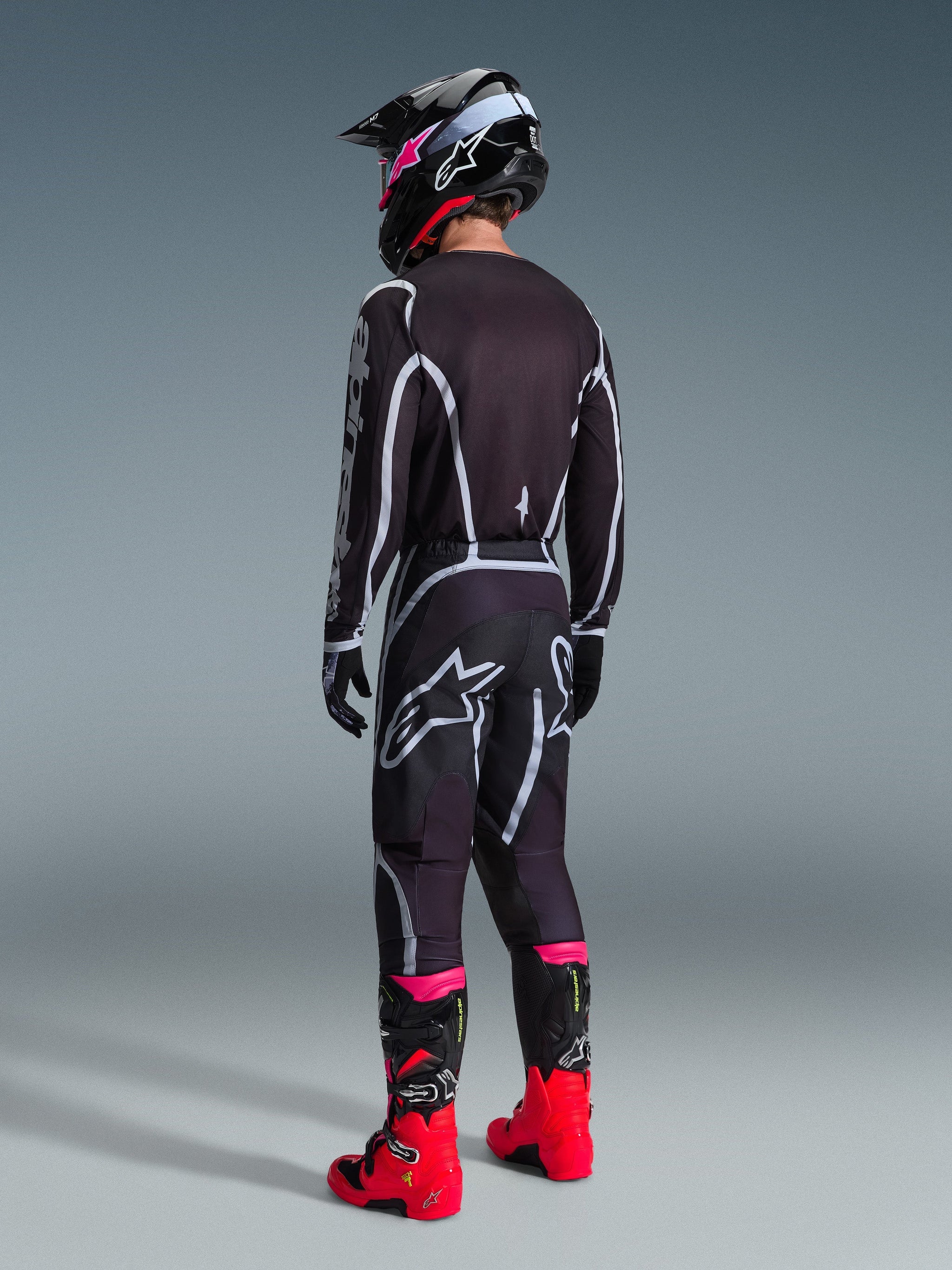 Motocross rider wearing complete racing gear, Black and Gray 2026 Fluid Apex Jersey seen from behind, matching pants with white and gray accents, black helmet, and vibrant red and pink boots, coordinated outfit for off-road motocross