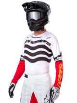 Motocross rider wearing the Alpinestars Limited Edition Techstar Deep Jersey, white jersey featuring black wavy stripes and red lower sleeves, styled with a black helmet, mirrored goggles, white and black gloves, and white racing pants, performance motocross outfit.