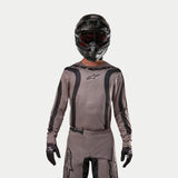 Motocross rider wearing the Alpinestars 2024 Fluid Lurv Jersey in Mud Black, an earthy grey-brown fabric with black graphic stripes and a star logo on the chest, paired with a black helmet, red goggles, and black gloves for a coordinated off-road racing look