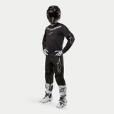 Motocross rider wearing Alpinestars 2024 Fluid Graphite Jersey in Black and Silver, matching black pants with silver star logos, white and black boots, white and black gloves, and a black helmet with red goggles, full off-road racing gear