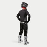 Motocross rider wearing the Alpinestars 2024 Fluid Graphite Jersey in Black Silver from a rear view, long-sleeve athletic fit jersey paired with black motocross pants, black helmet, and white and black boots, complete professional gear for off-road racing.