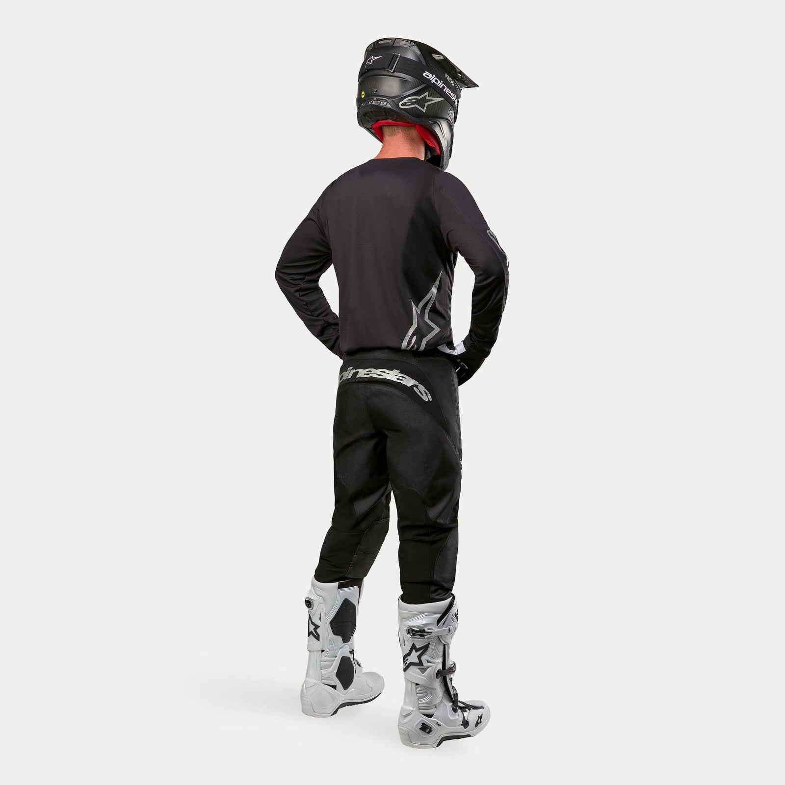 Motocross rider wearing the Alpinestars 2024 Fluid Graphite Jersey in Black Silver from a rear view, long-sleeve athletic fit jersey paired with black motocross pants, black helmet, and white and black boots, complete professional gear for off-road racing.