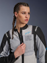 Woman wearing the Alpinestars Stella ST-2 Air Jacket in Ice Gray and Black, close-up portrait, featuring mesh panels for ventilation, 'ALPINESTARS EXPLORATION EQUIPMENT' branding on the chest with a blue logo, modern adventure motorcycle jacket