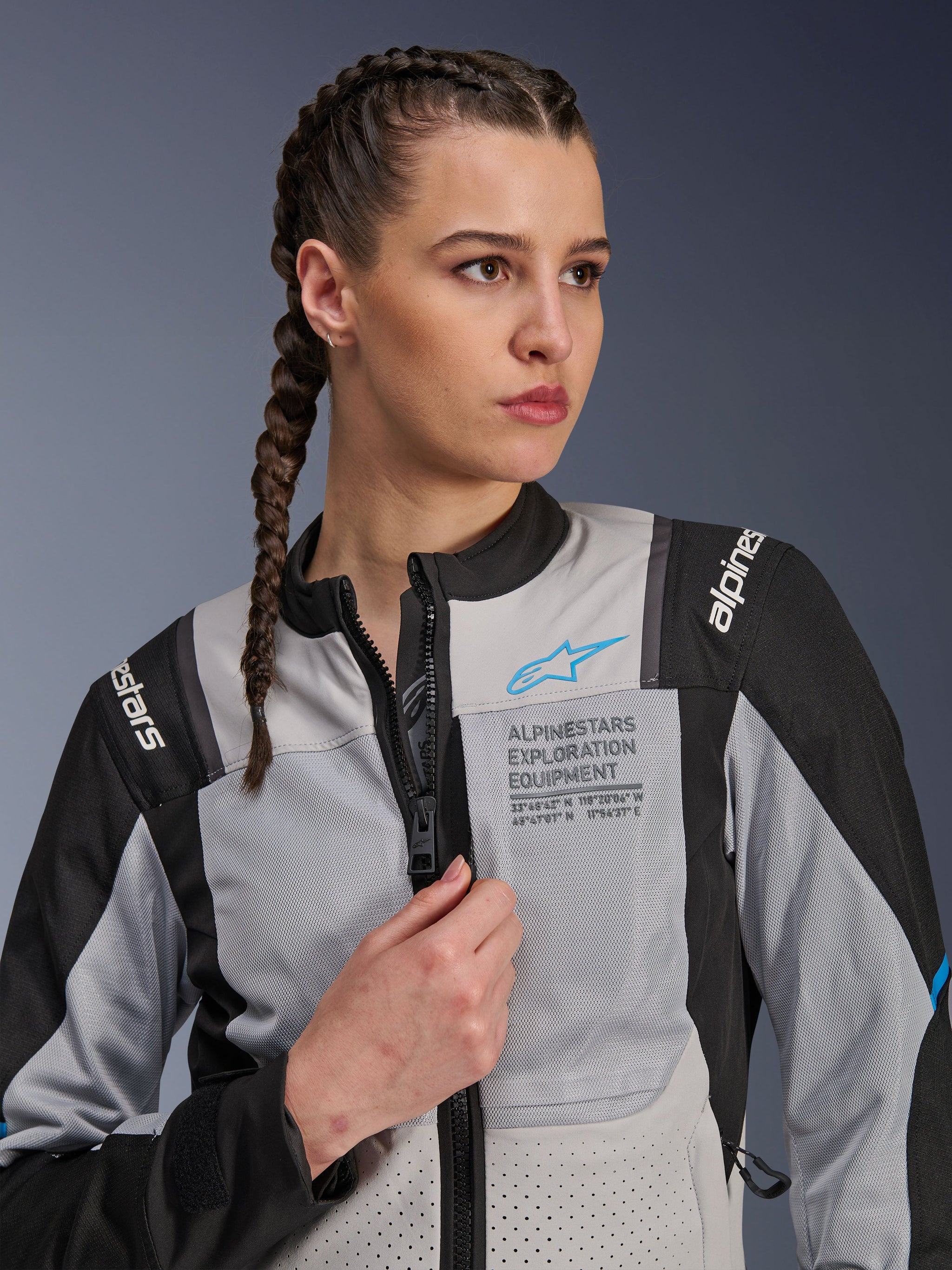 Woman wearing the Alpinestars Stella ST-2 Air Jacket in Ice Gray and Black, close-up portrait, featuring mesh panels for ventilation, 'ALPINESTARS EXPLORATION EQUIPMENT' branding on the chest with a blue logo, modern adventure motorcycle jacket