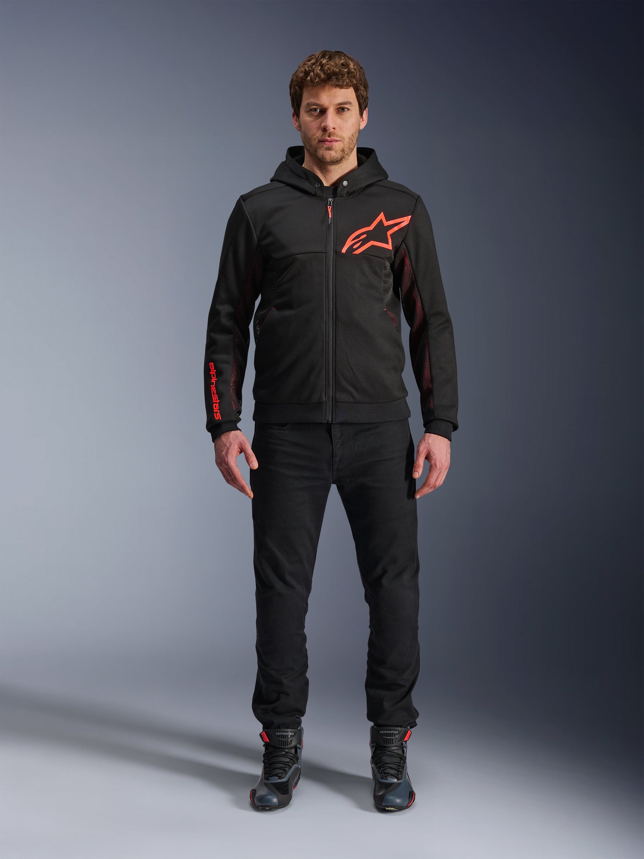 Alpinestars Chrome Air Sport Hoodie, Black and Red Fluo, Urban Motorcycle Riding Jacket with tech fleece construction, featuring a large red Astars logo on the chest and branding on the sleeve, worn by a model with black pants and riding shoes