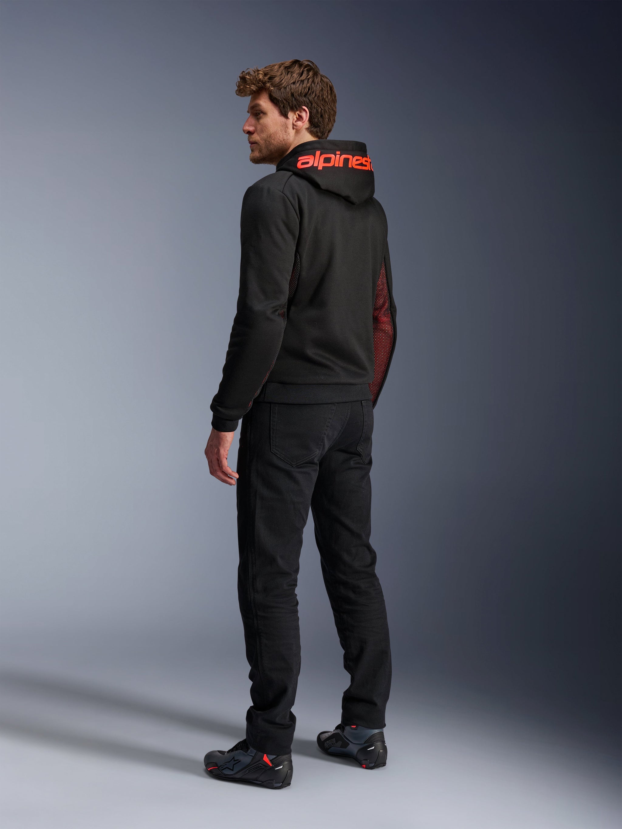 Alpinestars Chrome Air Sport Hoodie, urban motorcycle hoodie, Black and Red Fluo, rear view showing red Alpinestars branding on the hood and mesh side panels, worn with black riding pants and technical footwear