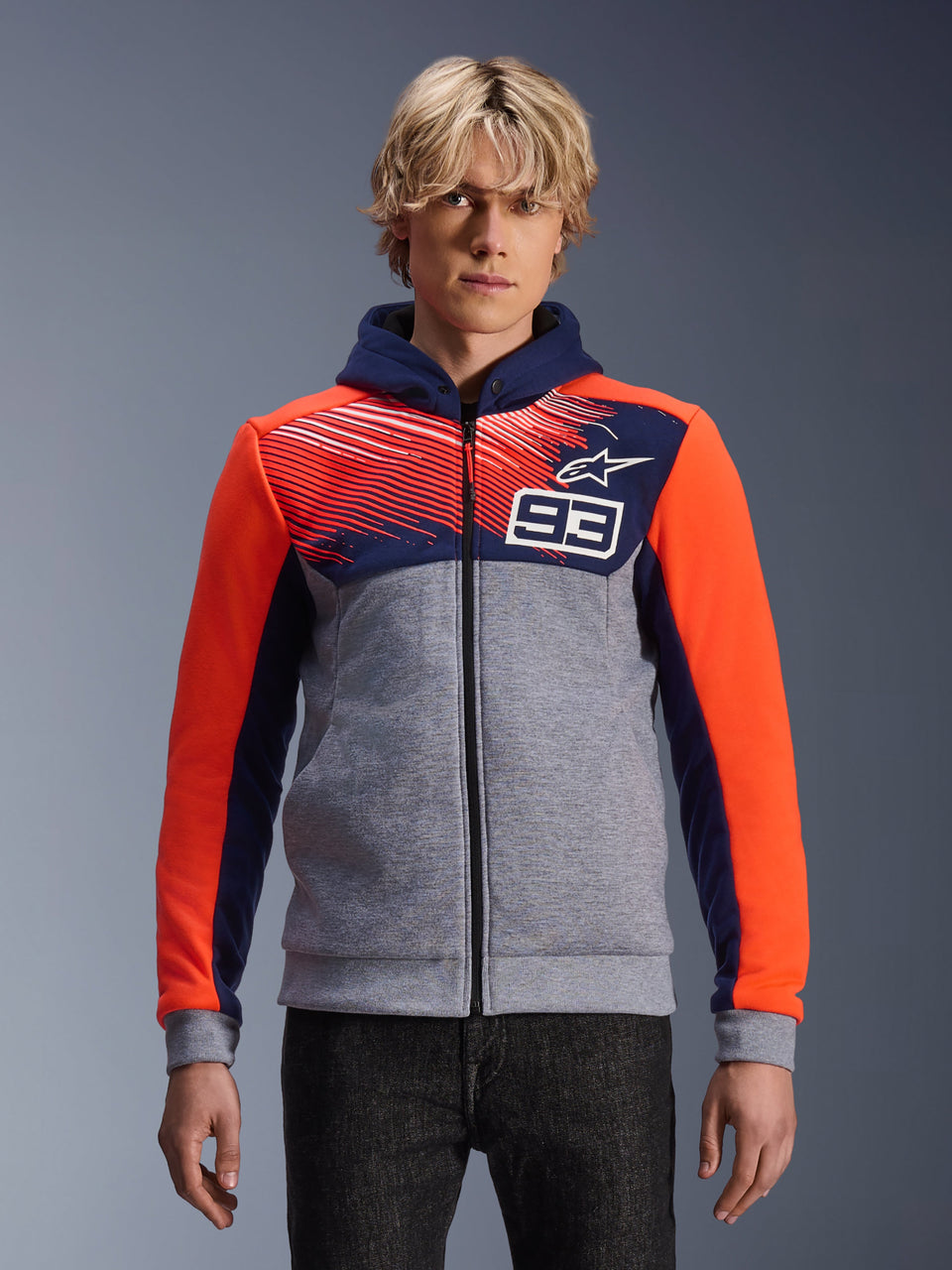 Alpinestars MM93 Chrome V2 Sport Hoodie, Motorcycle Zip-Up Hoodie, Gray melange with Navy Blue chest and hood, Bright Orange sleeves, Number 93 chest design with striped pattern and star logo, worn by model with dark denim, Marc Marquez Collection.