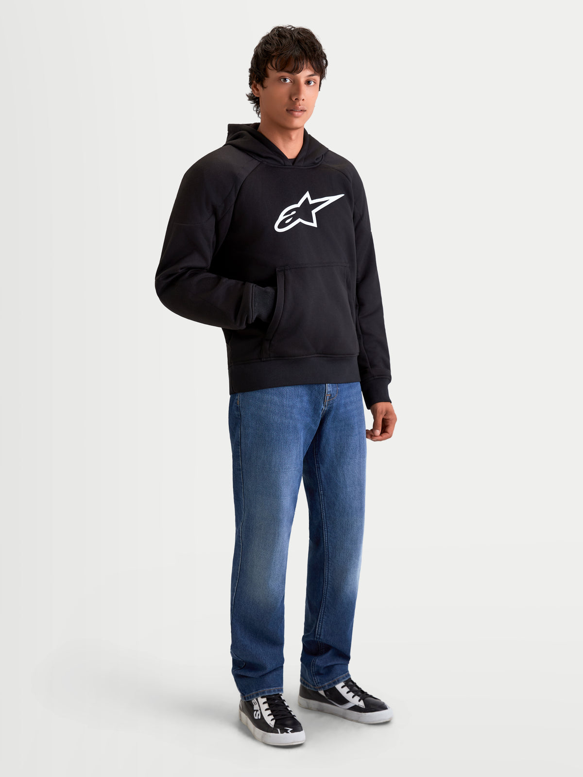 Alpinestars Ageless Hoodie, men's urban motorcycle riding hoodie in black, front view on a model wearing casual denim jeans, featuring a prominent white Astar logo on the chest and internal abrasion reinforcements.
