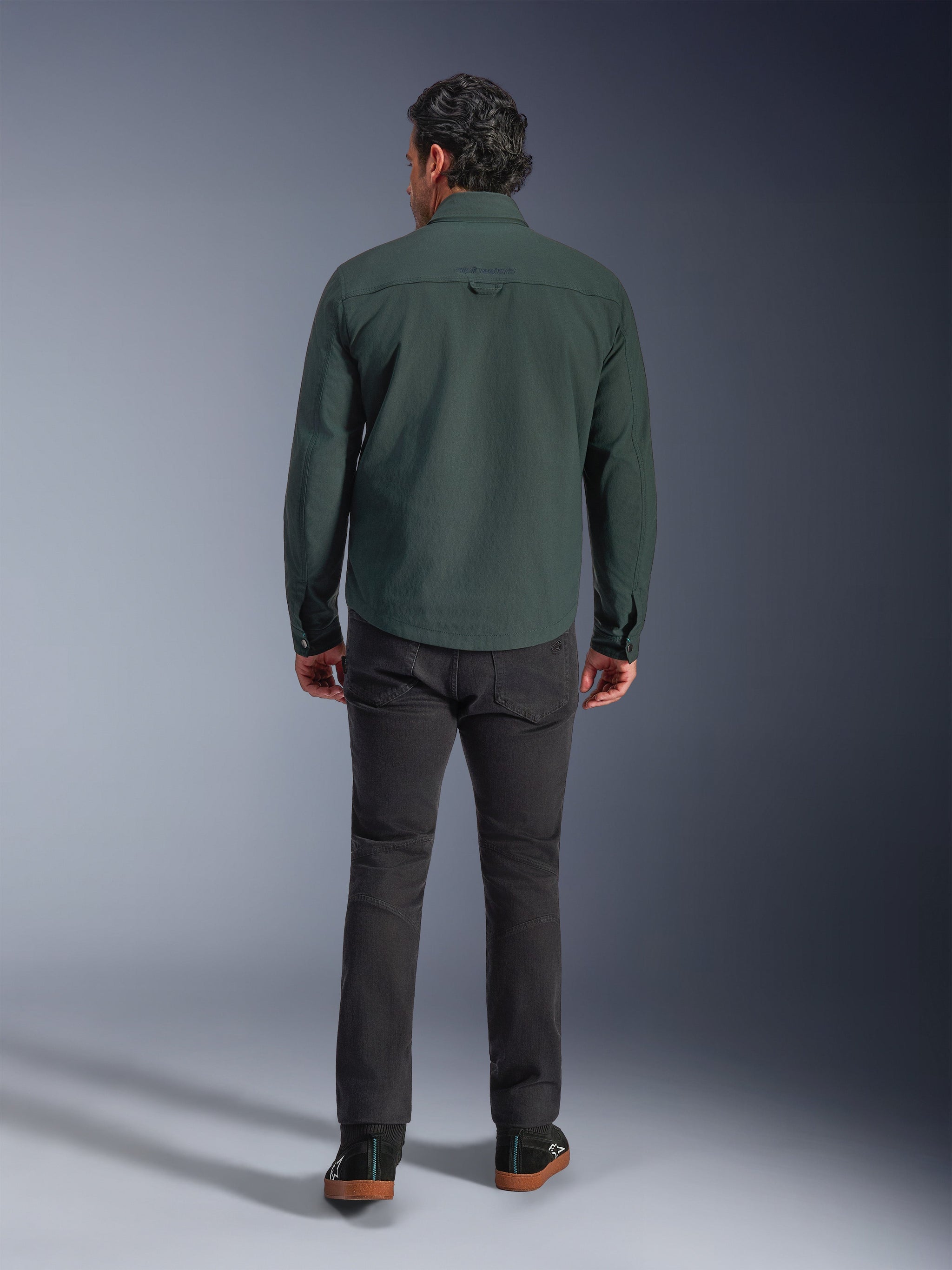 Alpinestars Bryon Water Resistant Overshirt in Dark Emerald, back view on model, paired with dark grey motorcycle pants and black riding shoes, showcasing a classic workwear-inspired design with a slightly longer back for riding coverage.