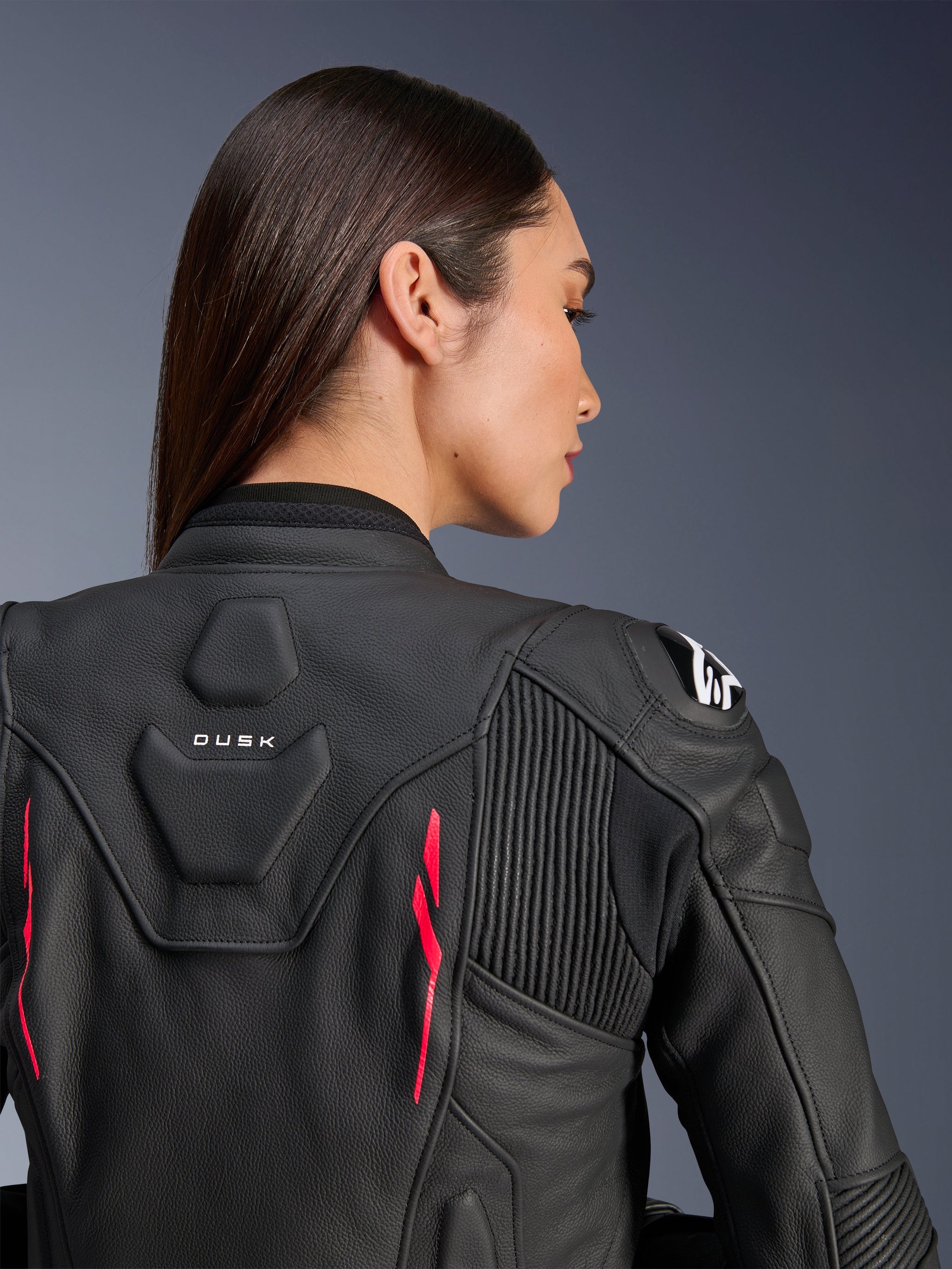 Woman wearing Alpinestars Stella Dusk Leather Jacket, black leather with Diva Pink accents and white logo on shoulder, back view showing DUSK branding and accordion stretch panels, sporty ergonomic design