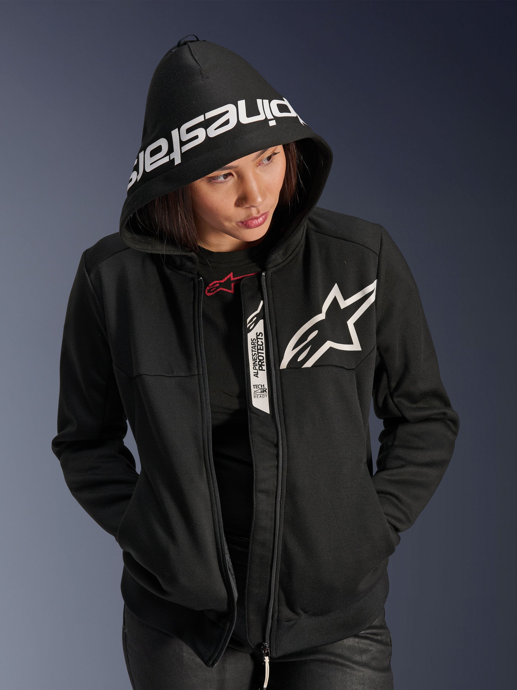 Alpinestars Woman Stella Chrome V2 Sport Hoodie, Black and White, motorcycle riding hoodie with a white star logo on the chest and Alpinestars branding on the hood brim, woman wearing hood up, casual urban sport style with Tech-Air Ready details.