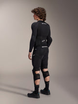 Model wearing Alpinestars A-Impact Plasma Pro Knee Shin Protectors from the back, Black and White, showing the triple strap closure and ventilated mesh panels, worn with a full set of black mountain bike protective gear including a long-sleeve base layer and padded shorts
