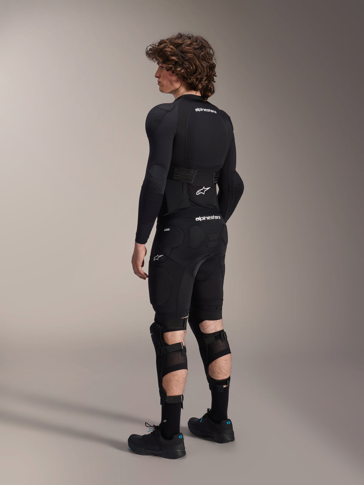 Model wearing Alpinestars A-Impact Plasma Pro Knee Shin Protectors from the back, Black and White, showing the triple strap closure and ventilated mesh panels, worn with a full set of black mountain bike protective gear including a long-sleeve base layer and padded shorts