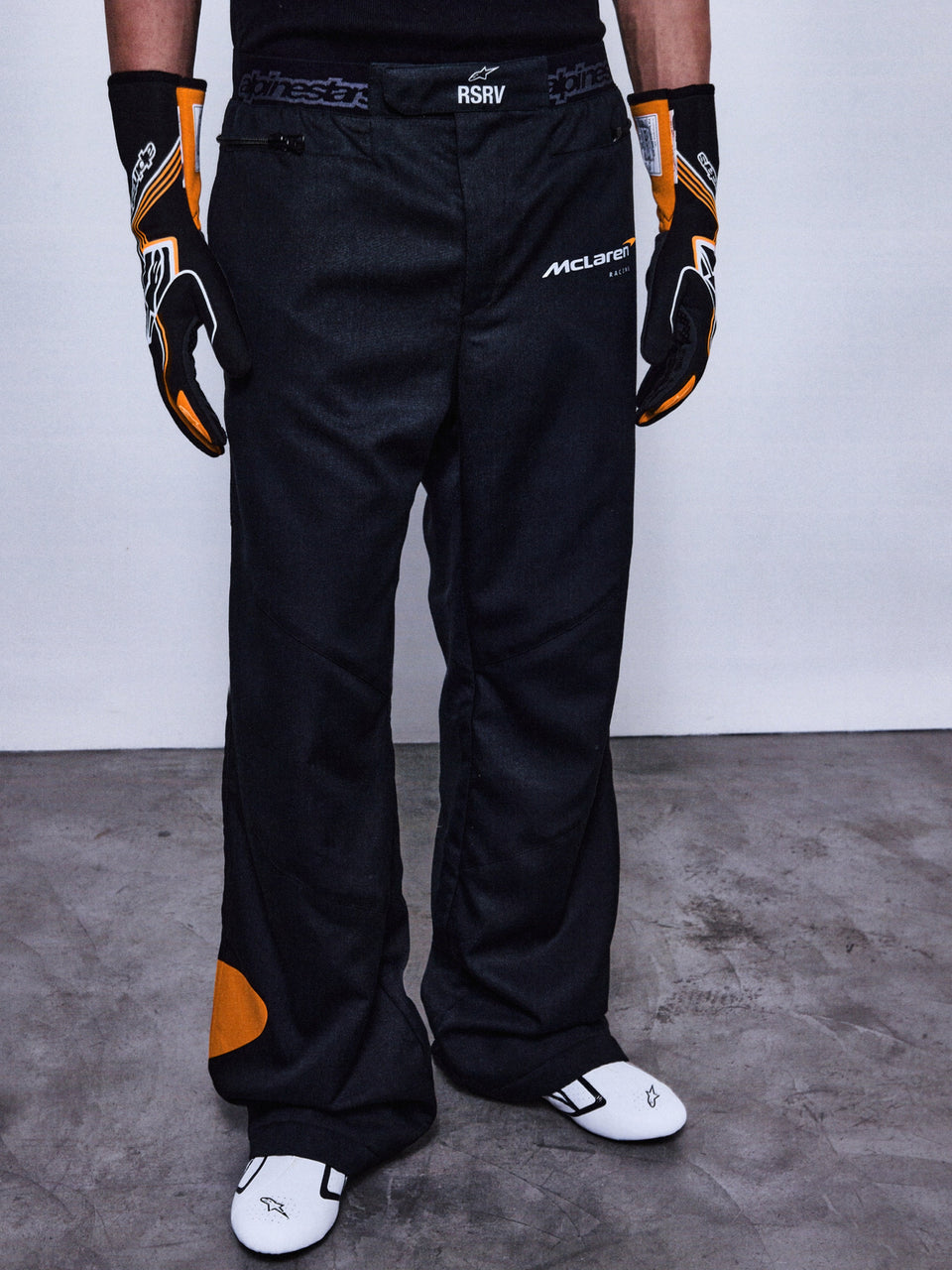 RSRV Arena Suit Pants, Black Racing Pants with McLaren and Alpinestars Logos, Orange Side Patches, Premium Motorsport Apparel, Worn with Black and Orange Gloves and White Racing Shoes