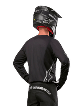 Motocross rider viewed from the back wearing the Alpinestars 2024 Fluid Graphite Jersey in Black Silver, featuring a large silver logo on the side, paired with a black carbon-texture helmet and black pants, professional off-road racing gear