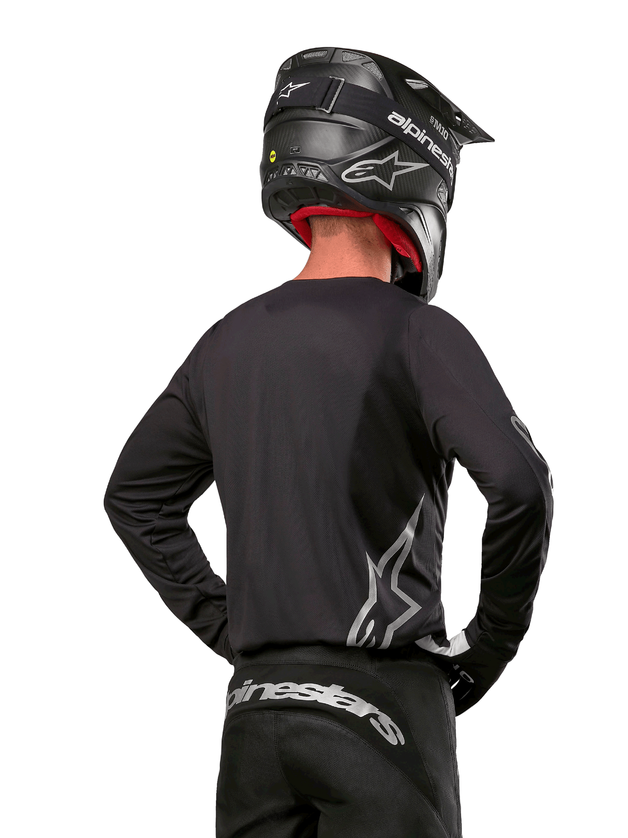 Motocross rider viewed from the back wearing the Alpinestars 2024 Fluid Graphite Jersey in Black Silver, featuring a large silver logo on the side, paired with a black carbon-texture helmet and black pants, professional off-road racing gear