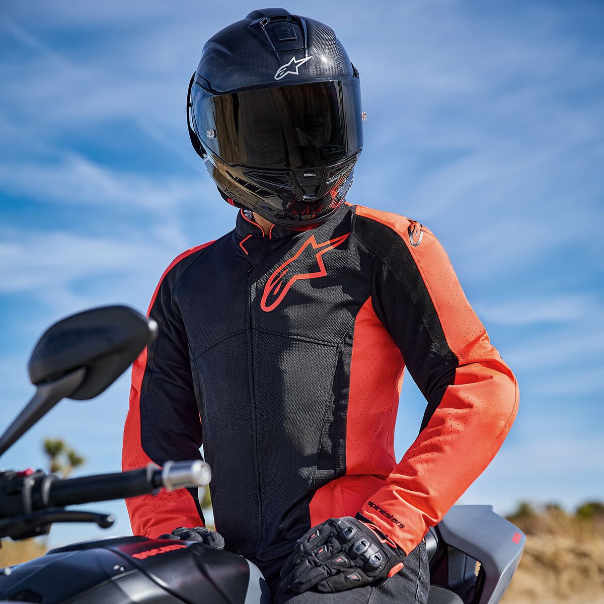 Alpinestars Viper Air V4 Jacket in Red Fluo and Black, worn by a rider on a motorcycle, featuring a large chest logo and ventilated mesh panels, paired with a carbon fiber helmet and black gloves, road and sport motorcycling gear