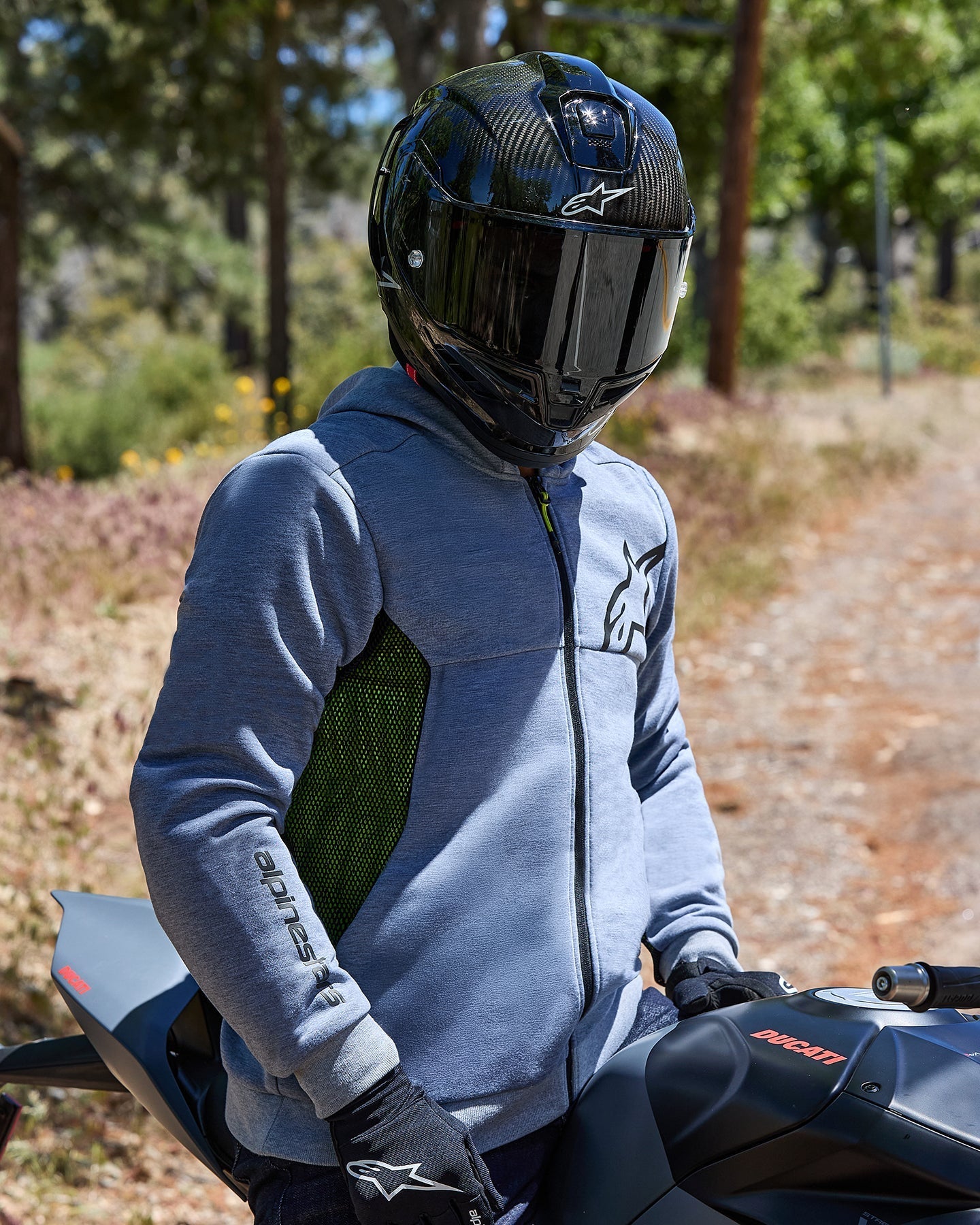 Motorcycle rider wearing the Alpinestars Chrome Air Sport Hoodie in Gray Heather, featuring lime green mesh side panels and black logos, paired with a black carbon fiber helmet and black gloves, seated on a Ducati bike in an outdoor road setting